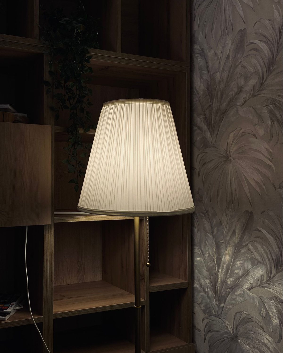 Botanical wallpaper behind dark wood shelving, lit softly by a floor lamp