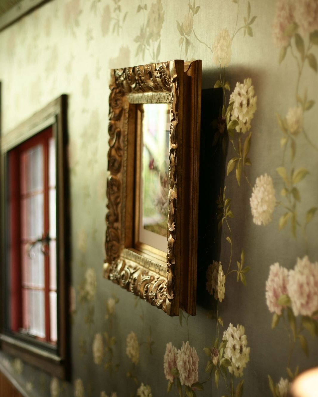 Green floral wallpaper with a gilded framed mirror and a soft table lamp